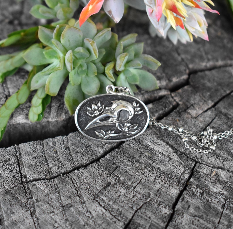 May include: A silver pendant necklace featuring a detailed, three-dimensional design of a crow skull with floral accents. The pendant is oval-shaped and hangs from a silver chain.