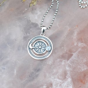 May include: A silver necklace with a circular pendant featuring a sun, moon, and a small blue stone. The pendant is detailed with a textured design.