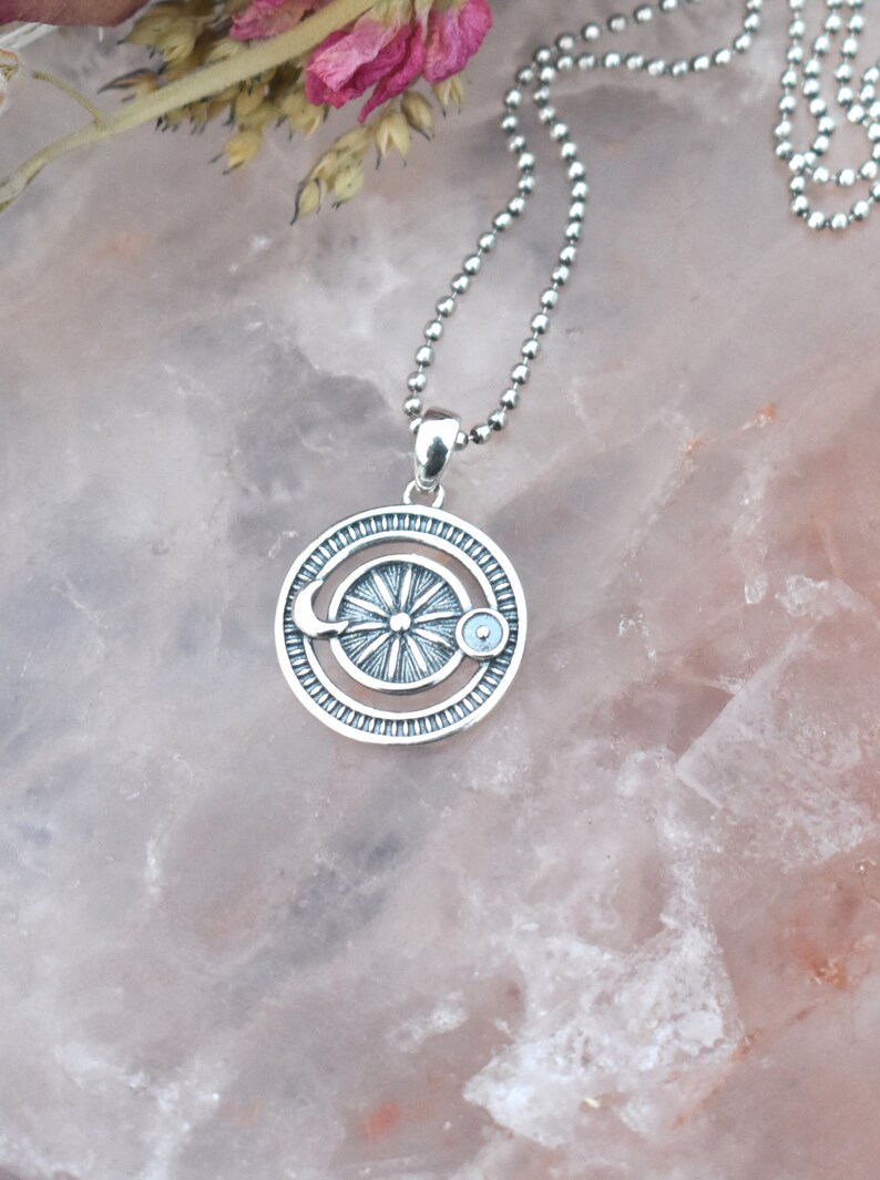 May include: A silver necklace with a circular pendant featuring a sun, moon, and a small blue stone. The pendant is detailed with a textured design.