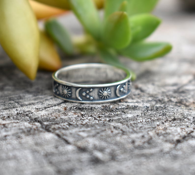 May include: A silver ring with a celestial design featuring suns, moons, and stars. The ring is on a wooden surface.