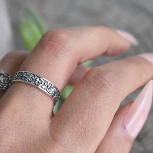 May include: A silver ring with a floral design. The ring is worn on a finger with light pink nail polish.