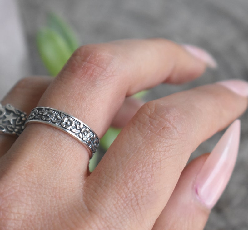 May include: A silver ring with a floral design. The ring is worn on a finger with light pink nail polish.