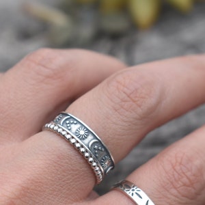 May include: A silver ring with a beaded band and a wider band featuring a repeating pattern of moons, stars, and suns.