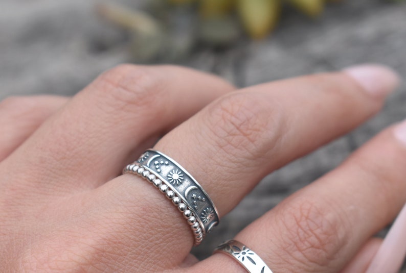 May include: A silver ring with a beaded band and a wider band featuring a repeating pattern of moons, stars, and suns.