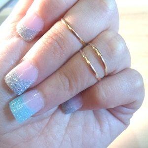May include: Close-up of a hand with gold rings and manicured nails. The nails feature a French manicure design with a pink base and glittery silver and blue tips. Two thin gold rings adorn the fingers.