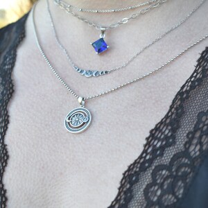 May include: A silver necklace with a layered chain design. The top chain features a square blue gemstone pendant. The bottom chain features a circular pendant with a sun and moon design.