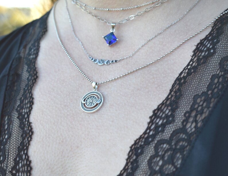May include: A silver necklace with a layered chain design. The top chain features a square blue gemstone pendant. The bottom chain features a circular pendant with a sun and moon design.