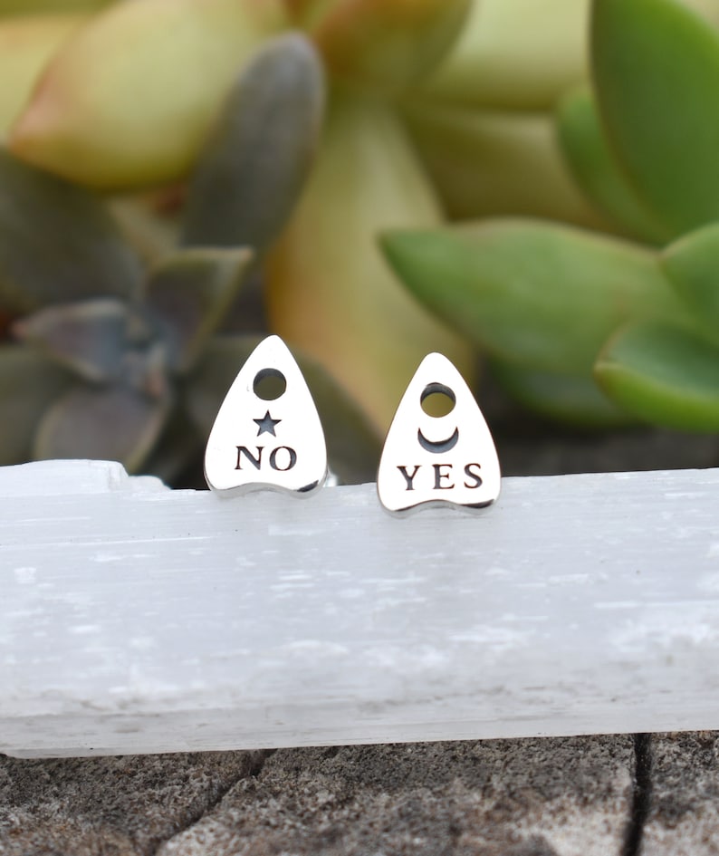 May include: A pair of silver earrings shaped like a Ouija planchette with the words "YES" and "NO" engraved on them. The earrings are on a white surface.