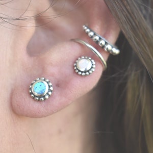 May include: A close-up of a person's ear with three silver earrings. The earrings are all round and have a beaded design. The earring in the lower lobe has a blue opal stone, the earring in the upper lobe has a white opal stone, and the earring in the helix has a silver hoop with a beaded design.