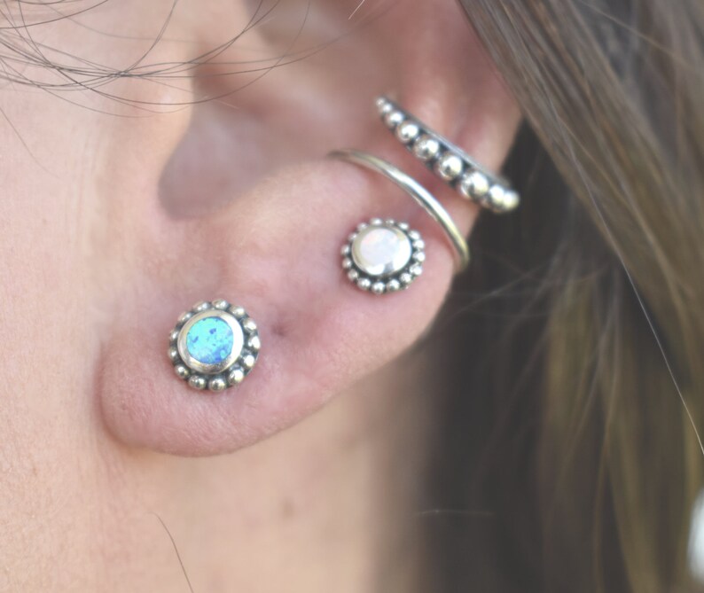 May include: A close-up of a person's ear with three silver earrings. The earrings are all round and have a beaded design. The earring in the lower lobe has a blue opal stone, the earring in the upper lobe has a white opal stone, and the earring in the helix has a silver hoop with a beaded design.