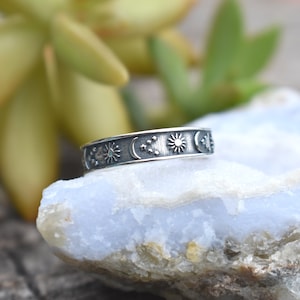 May include: A silver ring with a celestial design featuring a sun, moon, and stars. The ring is on a light blue rock.