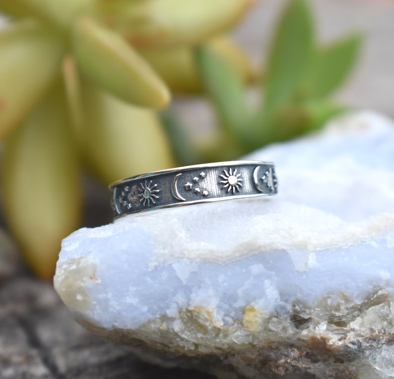 May include: A silver ring with a celestial design featuring a sun, moon, and stars. The ring is on a light blue rock.