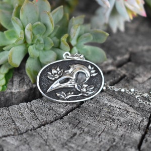 May include: Silver pendant necklace featuring a detailed raven skull with floral accents. The pendant is oval shaped and hangs from a silver chain.