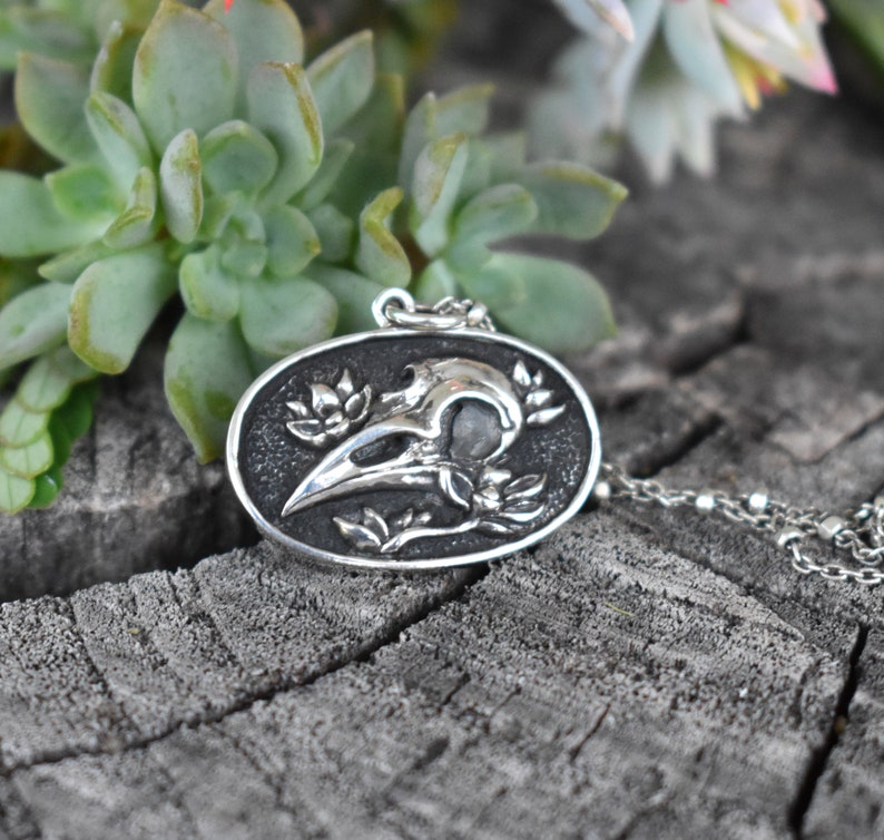 May include: Silver pendant necklace featuring a detailed raven skull with floral accents. The pendant is oval shaped and hangs from a silver chain.