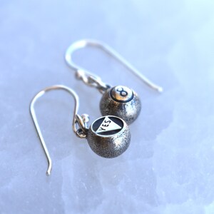 May include: A pair of silver earrings with a textured finish. One earring features a black triangle with the word "YES" inscribed inside, while the other earring features a black 8-ball.
