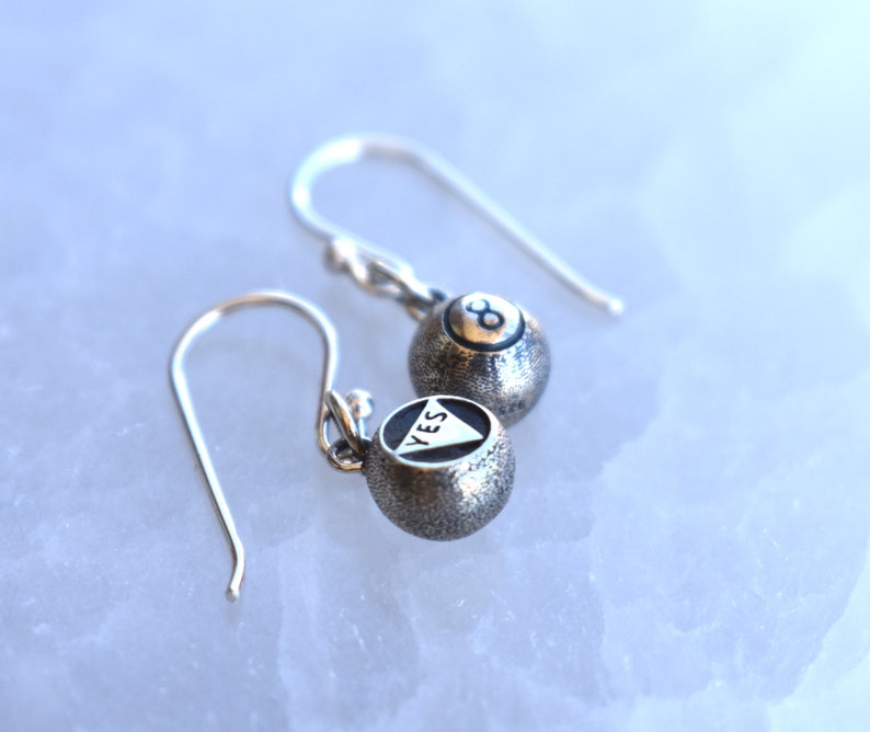 May include: A pair of silver earrings with a textured finish. One earring features a black triangle with the word "YES" inscribed inside, while the other earring features a black 8-ball.