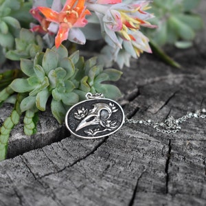 May include: A silver pendant necklace featuring a detailed crow skull with floral accents. The pendant is oval-shaped and hangs from a silver chain. The necklace is displayed on a wooden surface with green succulents.