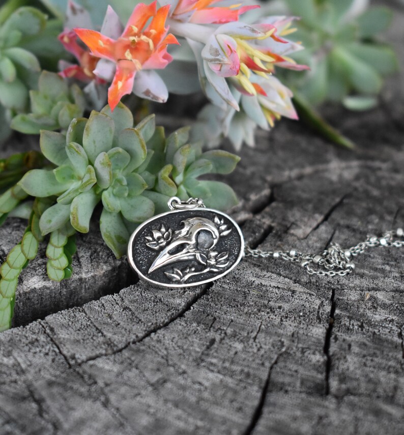 May include: A silver pendant necklace featuring a detailed crow skull with floral accents. The pendant is oval-shaped and hangs from a silver chain. The necklace is displayed on a wooden surface with green succulents.