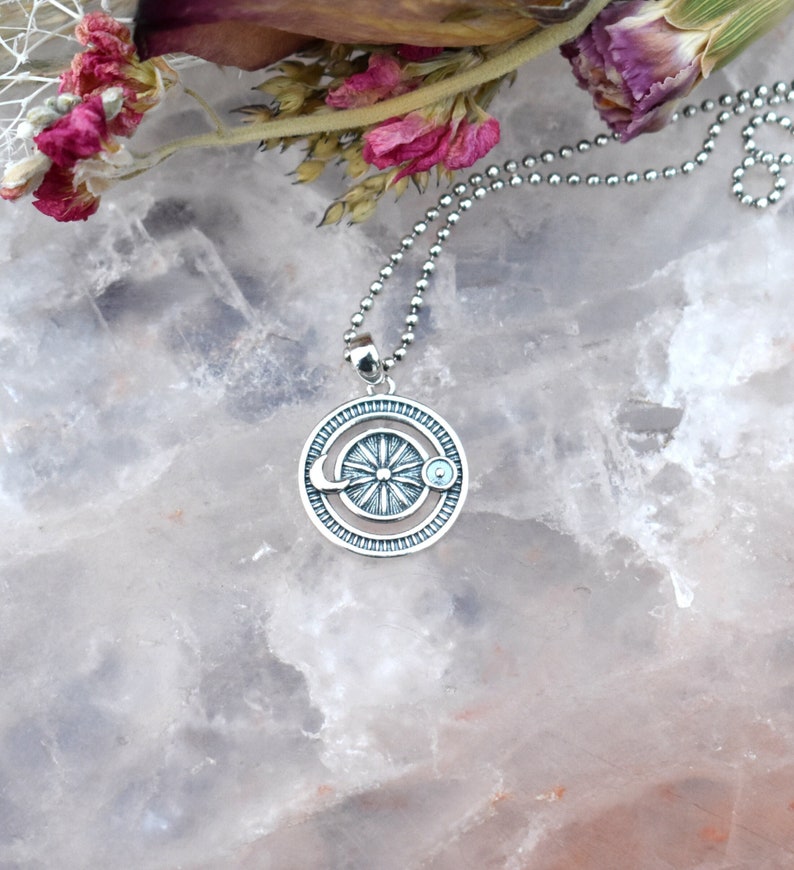 May include: A silver necklace with a circular pendant featuring a sun, moon, and a small blue gemstone. The pendant is detailed with a textured design.