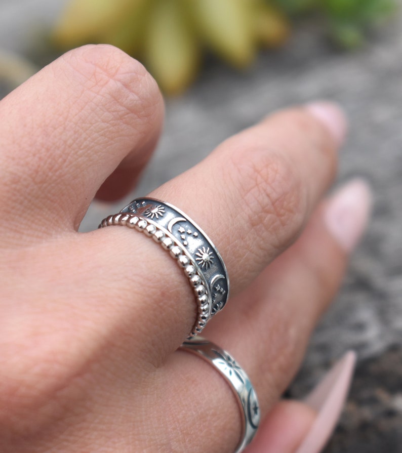 May include: A silver ring with a design of moons, stars, and suns. The ring is worn on a finger.