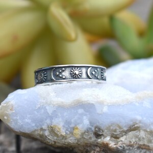 May include: A silver ring with a celestial design featuring a sun, moon, and stars. The ring is on a light blue rock.