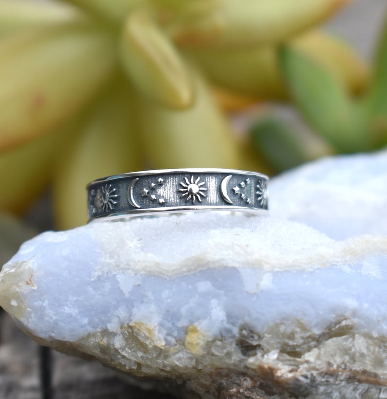 May include: A silver ring with a celestial design featuring a sun, moon, and stars. The ring is on a light blue rock.