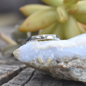 May include: A silver ring with a sunburst design. The ring is on a light blue and white rock.