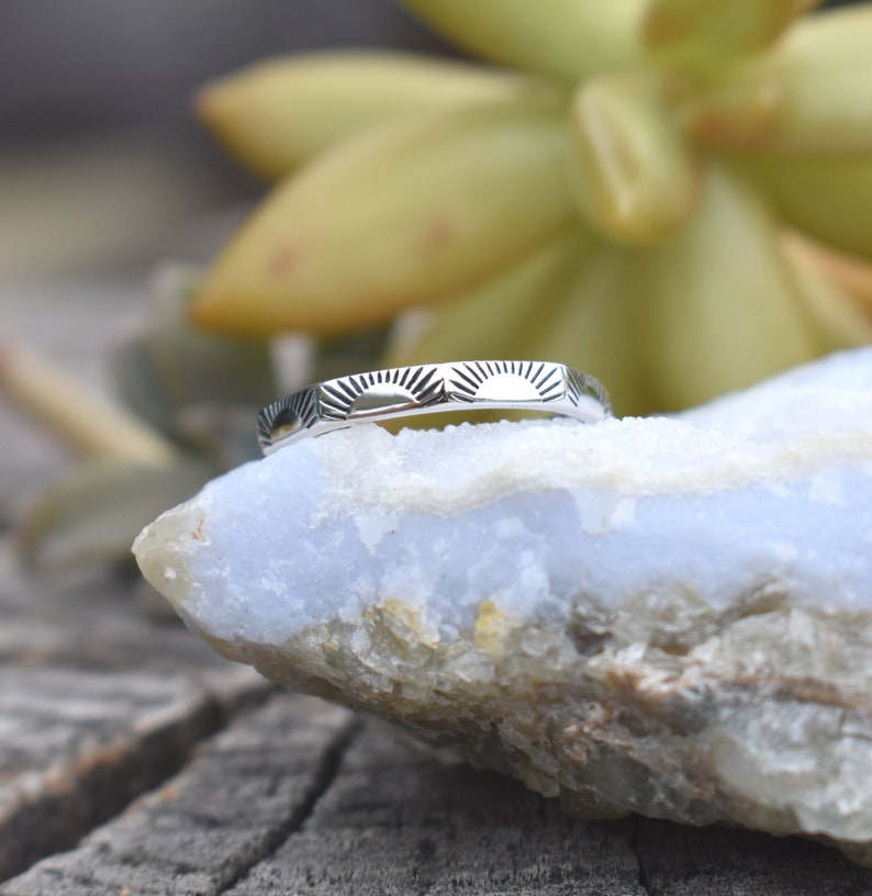 May include: A silver ring with a sunburst design. The ring is on a light blue and white rock.