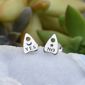 May include: A pair of silver earrings shaped like a Ouija board planchette. One earring has a crescent moon and the word "YES" engraved on it. The other earring has a star and the word "NO" engraved on it.