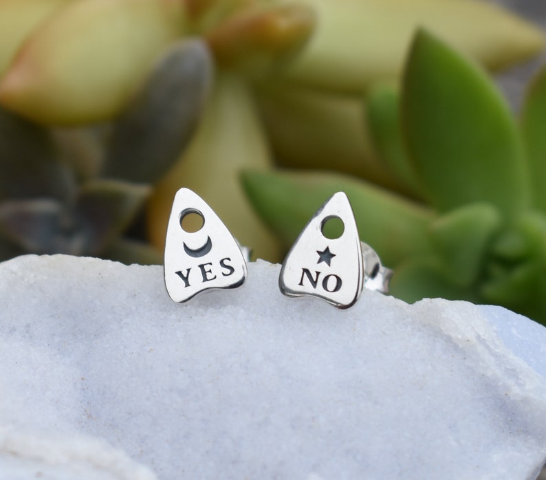 May include: A pair of silver earrings shaped like a Ouija board planchette. One earring has a crescent moon and the word "YES" engraved on it. The other earring has a star and the word "NO" engraved on it.