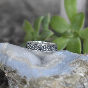 May include: A silver band ring with a floral design. The ring is on a light blue and gray rock.