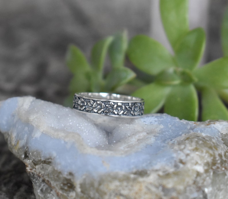 May include: A silver band ring with a floral design. The ring is on a light blue and gray rock.