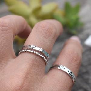 May include: Two silver rings on a finger. The top ring has a sun design and the bottom ring has a beaded design. The ring on the other finger has a star and moon design.