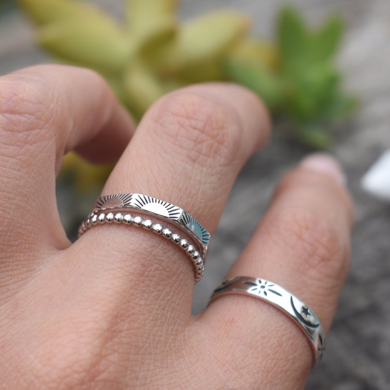 May include: Two silver rings on a finger. The top ring has a sun design and the bottom ring has a beaded design. The ring on the other finger has a star and moon design.