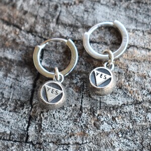 May include: A pair of silver hoop earrings with a small charm hanging from each hoop. The charms are round and have the word "YES" inscribed on them.