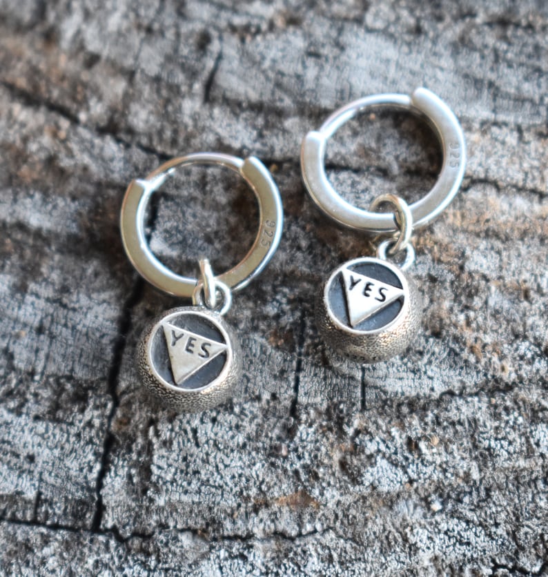 May include: A pair of silver hoop earrings with a small charm hanging from each hoop. The charms are round and have the word "YES" inscribed on them.
