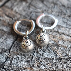 May include: A pair of silver hoop earrings with a silver 8-ball charm hanging from each hoop.