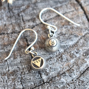 May include: A pair of silver earrings with a small charm on each. One charm is a silver ball with the number 8 on it. The other charm is a silver ball with the word "YES" on it.