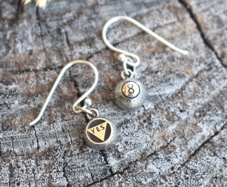 May include: A pair of silver earrings with a small charm on each. One charm is a silver ball with the number 8 on it. The other charm is a silver ball with the word "YES" on it.
