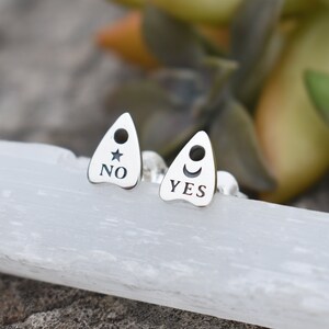 May include: A pair of silver earrings shaped like a Ouija board planchette. One earring says "YES" with a crescent moon and the other says "NO" with a star.