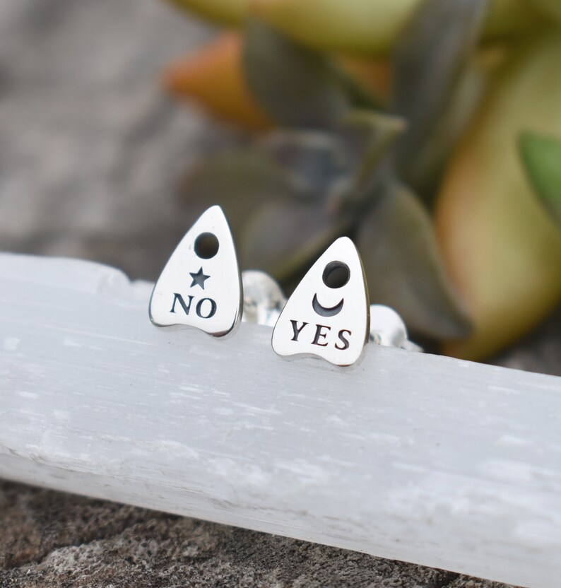 May include: A pair of silver earrings shaped like a Ouija board planchette. One earring says "YES" with a crescent moon and the other says "NO" with a star.