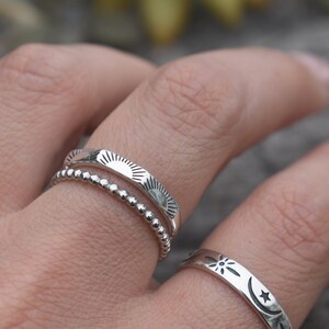 May include: Three silver rings with different designs. The top ring has a sun design, the middle ring has a beaded design, and the bottom ring has a moon and star design.