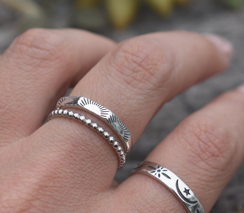 May include: Three silver rings with different designs. The top ring has a sun design, the middle ring has a beaded design, and the bottom ring has a moon and star design.