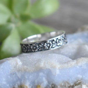 May include: A silver band ring with a floral design. The ring is on a light blue rock.