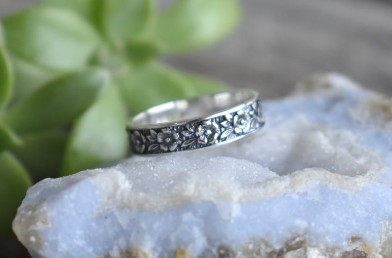 May include: A silver band ring with a floral design. The ring is on a light blue rock.