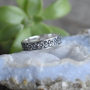 May include: A silver band ring with a floral design. The ring is on a light blue background.