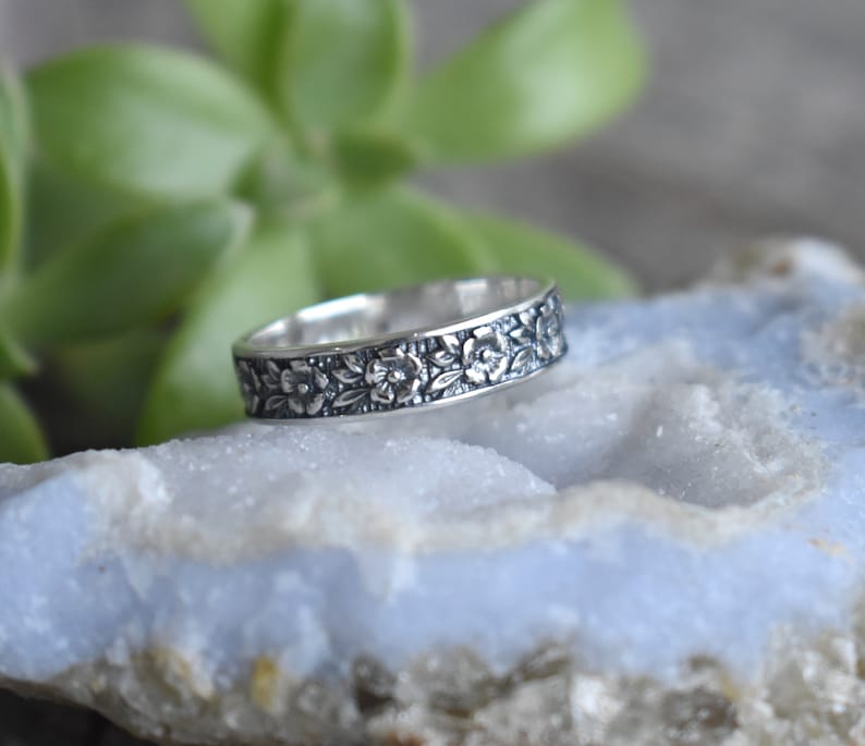 May include: A silver band ring with a floral design. The ring is on a light blue background.