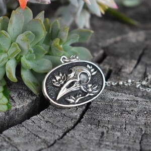 May include: Silver pendant necklace featuring a detailed skull of a crow with floral accents. The pendant is oval shaped and has a black background.