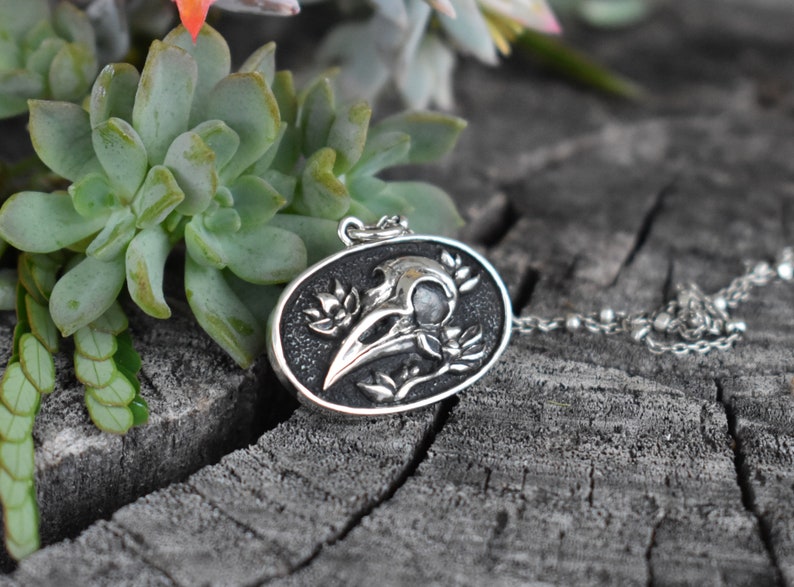 May include: Silver pendant necklace featuring a detailed skull of a crow with floral accents. The pendant is oval shaped and has a black background.