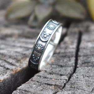 May include: A silver ring with a celestial design featuring moons, stars, and suns. The ring is on a wooden surface.